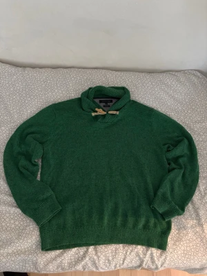 Grön stickad tröja Tommy Hilfiger - Snygg grön stickad tröja från Tommy Hilfiger med grovstickad krage och dekorativt snöre med trädetalj. Tillverkad i en mix av linne och bomull för en skön känsla. Perfekt för dig som vill ha en unik och bekväm tröja med coola detaljer.