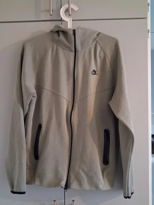 Grå hoodie från Nike med dragkedja - Säljer en stilren grå hoodie från Nike tech med svart dragkedja och svarta detaljer vid fickorna. Hoodien har huva och ett diskret Nike-logo på bröstet. Perfekt för dig som gillar sportig och enkel design.