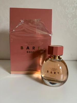 Victoria's Secret Bare Rose parfym 100ml  - En elegant parfym från Victoria's Secret, Bare Rose. Flaskan är rund med roséguldigt lock och detaljer, och doften presenteras i en stilren rosa förpackning med gulddetaljer. Perfekt för dig som gillar fräscha och blommiga toner.