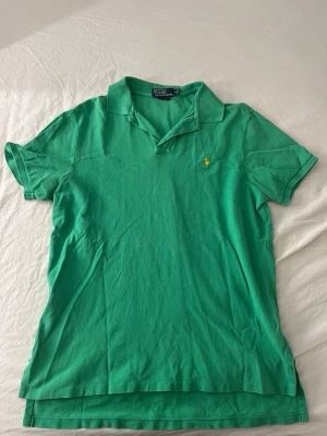 Grön pikétopp från Polo Ralph Lauren - Grön pikétopp från Polo Ralph Lauren med klassisk krage, korta ärmar och gul broderad logga på bröstet. Tillverkad i mjuk bomull för en bekväm och sportig stil.