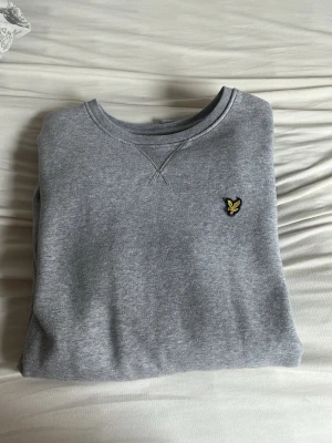 Grå sweatshirt från Lyle & Scott - Klassisk grå sweatshirt från Lyle & Scott med rund halsringning och den ikoniska gula örnloggan broderad på bröstet. Tröjan har en avslappnad passform och är tillverkad i mjuk bomullsblandning. Perfekt för en casual och stilren look.