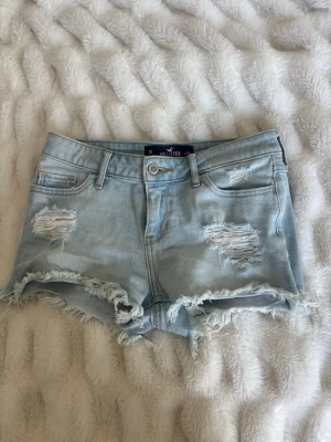  jeansshorts Hollister - Säljer ett par ljusblå jeansshorts från Hollister med låg midja och slitna detaljer. Shortsen har fransiga benslut och klassisk femficksdesign. Perfekta för sommaren och ger en avslappnad vibe.
