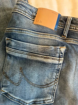 Ljusblå lowrise bootcut jeans från LTB - Säljer ett par ljusblå jeans från LTB med bootcut ben och klassisk femficksdesign. Jeansen har snygga slitningar och kontrastsömmar samt en läderpatch med logga bak i midjan. Perfekta för en avslappnad och trendig look.