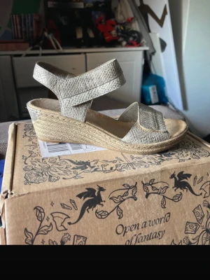 Beiga kilklackade sandaler - Snygga sandaler med kilklack och öppen tå i beige färg. Ovandelen är i texturerat material och klacken är klädd i flätad jute för en somrig känsla. Kardborreband runt vristen gör dem enkla att ta på och av. Perfekta för varma dagar.
