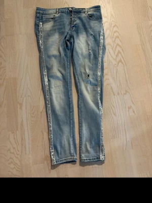 Blå jeans med silverrevärer - Snygga blå jeans med slitningar och silverfärgade revärer längs båda sidorna. Jeansen har normal midja, fem fickor och stängs med dubbla knappar. Perfekta för dig som vill ha en trendig och unik look.