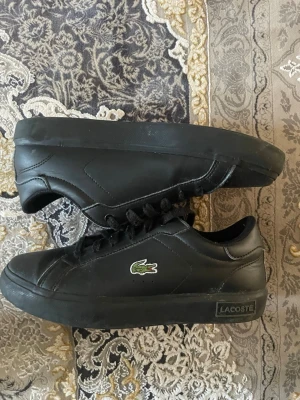 Svarta Lacoste sneakers i läder - Svarta sneakers från Lacoste i läder med klassisk låg profil. Skorna har svart sula, svarta skosnören och den ikoniska gröna krokodilloggan på sidan. Diskret Lacoste-märke på hälen och under sulan. Perfekt för en stilren och sportig look.