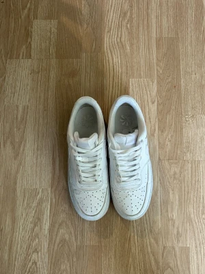 Vita Nike Air Force 1 sneakers - Klassiska vita Nike Air Force 1 sneakers med lågt skaft och perforerad tå. Skorna har en chunky sula och är tillverkade i läder med textildetaljer. Perfekta för en clean och stilren look.