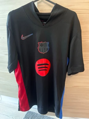 FC Barcelona matchtröja svart Nike - Snygg svart FC Barcelona matchtröja från Nike med röd och blå detalj på sidorna. Tryck med Spotify-logga på bröstet och klubbmärke. På ryggen står det 'Lamine Yamal' och nummer 19 i rött. Perfekt för dig som älskar fotboll och Barca. Storlek S/M