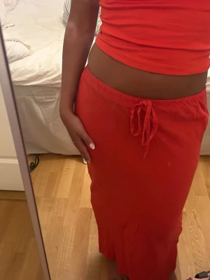 Röd långkjol med knyt i midjan - Säljer en röd långkjol med knyt i midjan. Kjolen har en enkel och avslappnad passform, perfekt för dig som gillar en chill och stilren look. Materialet känns mjukt och lätt, vilket gör den skön att bära under varma dagar.