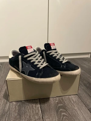 Mörk blå Golden goose SuperStars - Snygga Mörk blåa sneakers från Golden Goose med grå stjärndetalj på sidan, vita sulor och grå skosnören. Ovandelen är i mocka och skon har en vit hälkappa. Perfekta för en mer laidback stil. Passar perfekt nu till sommarn med lite varmare väder ute. Skorna är i väldigt bra skick utan några defekter. Skorna tvättas innan de skickas såklart. Box och dustbag tillkommer vid köp av skorna