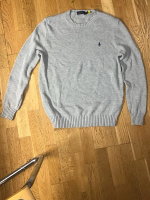 Ljusgrå stickad tröja från Polo Ralph Lauren - Snygg ljusgrå stickad tröja från Polo Ralph Lauren med klassisk blå logga broderad på bröstet. Tröjan har rund halsringning, långa ärmar och ribbade muddar vid ärmslut och nederkant. Perfekt för lager på lager och enkel att matcha.