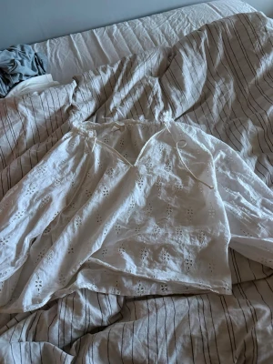 Vit broderad blus med spetsdetaljer - Säljer en vit blus med broderade hålmönster och romantiska spetsdetaljer. Blusen har tunna axelband och vida ärmar, perfekt för en luftig och somrig look. Materialet känns lätt och behagligt mot huden.