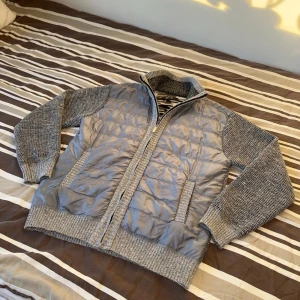Grå cardiganjacka med stickade ärmar - Snygg grå cardiganjacka med quiltad framsida och stickade ärmar samt rygg. Jackan har dragkedja, ståkrage och två fickor fram. Materialmix av vadderad polyester och mjuk stickad bomull. Perfekt för lager på lager och chill stil.