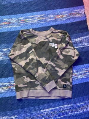 Camo sweatshirt med UG-logga - Säljer en camouflagemönstrad sweatshirt med UG-logga på bröstet. Tröjan har rund hals, ribbade muddar vid ärmslut och nederkant samt en relaxed fit. Perfekt för dig som gillar streetwear och vill sticka ut lite extra.