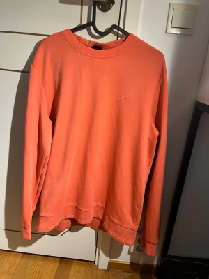 Korallfärgad sweatshirt från Hugo Boss - Säljer en stilren korallfärgad sweatshirt från Hugo Boss med diskret broderad logga på bröstet. Tröjan har rund halsringning, långa ärmar och ribbade muddar. Materialet är mjuk bomull och nederkanten har subtila Boss-detaljer.