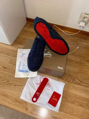 Mörkblå Louboutin sneakers - Snygga mörkblå sneakers från Christian Louboutin med klassisk röd sula och diskret logga. Skorna är tillverkade i mocka och har en låg profil med svarta snören. Perfekt för dig som vill sticka ut med exklusiva detaljer och lyxig känsla.