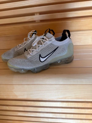 Nike Air Vapormax vita sneakers - Nike Air Vapormax sneakers i vitt och grått med svart Swoosh-logga på sidan. Ovandelen är i stickad mesh för extra andningsförmåga och sulan är genomskinlig med synliga luftbubblor för maxad komfort. Skorna har vita snören och svarta detaljer vid hälen.