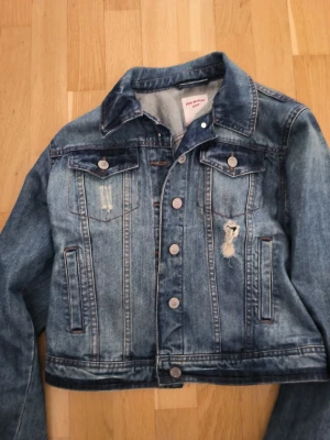 Blå jeansjacka från Pink Woman - Snygg blå jeansjacka från Pink Woman i klassisk modell med slitna detaljer och silverfärgade knappar. Två bröstfickor med lock och knappar samt två sidofickor. Jackan har en cool, lite croppad passform och markerade sömmar.