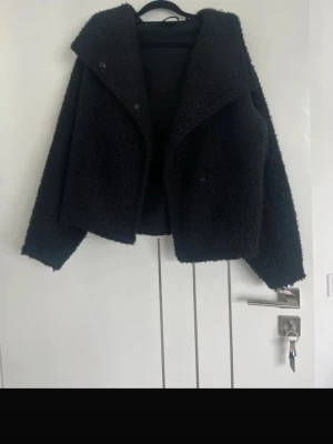 Svart teddyjacka med bred krage - Mysig svart teddyjacka med bred krage och knäppning framtill. Jackan har en boxig passform och är gjord i fluffigt teddy-material som ger en trendig look. Perfekt för kyliga dagar när du vill vara både varm och snygg.
