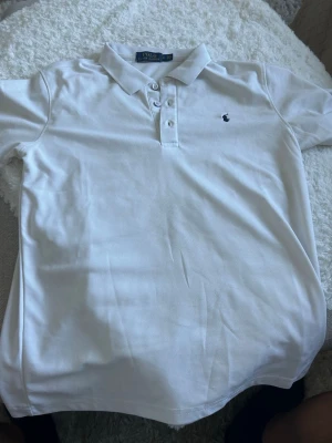 Vit pikétröja från Polo Ralph Lauren - Klassisk vit pikétröja från Polo Ralph Lauren med krage och knappar framtill. Tröjan har korta ärmar och den ikoniska broderade loggan på bröstet. Perfekt för en clean och stilren look.