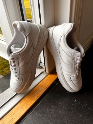 Vita klassiska sneakers i läder - Snygga helvita sneakers med lågt skaft och stilren design. Skorna har ovandel i läder, vita snören och diskreta sömmar för en clean look. Perfekta för dig som gillar minimalistisk stil och vill ha ett par fräscha sneakers till vardags.