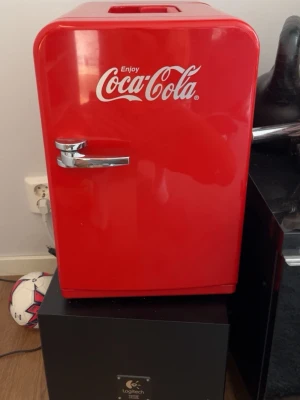 Röd Coca-Cola minikylskåp - Säljer en rött minikylskåp med Coca-Cola-logga. Kylen har en klassisk retrodesign med kromad handtag och vit insida med hylla. Perfekt för att förvara drycker eller snacks i rummet eller på kontoret. Tillverkad i plast och metall.