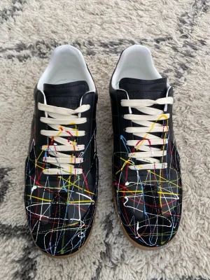 Maison margiela skor  - Unika svarta sneakers i läder med vita skosnören och färgstänk i rött, gult, blått och vitt över hela ovandelen. Snygg kontrast mellan den svarta basen och de livliga färgerna. Yttersulan är i gummi och insidan är vit. Perfekta för dig som vill sticka ut.