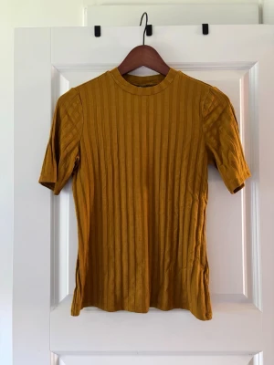 Ribbad senapsgul topp från Gina Tricot - Senapsgul ribbad topp från Gina Tricot med korta ärmar och rund hals. Toppen har en slim passform och vertikala ränder i materialet som ger en snygg struktur. Perfekt att styla med jeans eller kjol för en trendig look.