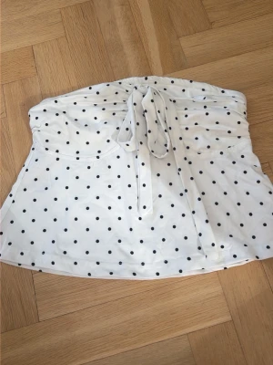 Vit prickig tubtopp med knyt - Snygg vit tubtopp med svarta prickar och knyt framtill. Toppen är axelbandslös och har en söt bandeau-stil som passar perfekt till sommaren. Materialet känns mjukt och lätt, perfekt för varma dagar.
