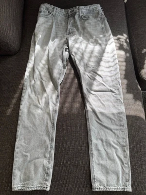 Ljusgrå raka jeans - Säljer ett par ljusgrå jeans med rak passform och klassisk femficksdesign. Jeansen har normal midja och är tillverkade i denim. Perfekta för en avslappnad och stilren look. Det är storlek 30/34 på jeansen.
