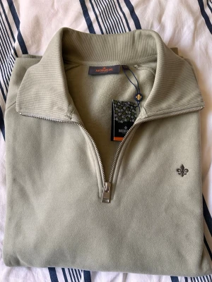 Olivgrön halfzip från Morris - Snygg olivgrön tröja från Morris med halv dragkedja och hög krage. Tröjan har en diskret logga broderad på bröstet och ribbade detaljer vid krage och ärmslut. Perfekt för en stilren och avslappnad look.  Model: Maryon, halfzip.     Helt oanvänd. Lappen kvar.             Ny pris 1 700 kr, pris kan diskuteras 