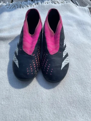 Adidas svarta och rosa fotbollsskor - Säljer ett par Adidas fotbollsskor i svart och rosa med vita detaljer och klassiska ränder på sidorna. Skorna har en strumpliknande passform utan snörning och är designade för bra bollkontroll. Perfekta för dig som vill sticka ut på planen.