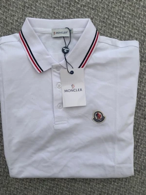 Vit Moncler pikétröja med logga - Vit pikétröja från Moncler med klassisk krage och knäppning framtill. Kragen har snygga ränder i rött och svart. Monclers ikoniska logga är broderad på bröstet. Perfekt för en clean och sportig look.