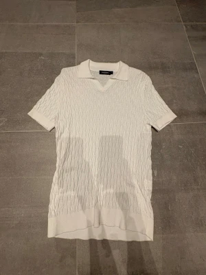 Vit stickad pikétröja Dressmann - Snygg vit stickad pikétröja från Dressmann med korta ärmar och klassisk krage. Tröjan har ett diskret kabelstickat mönster och ribbade muddar vid ärmslut och nederkant. Perfekt för en stilren och avslappnad look.