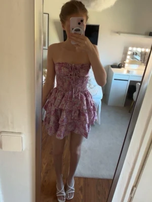 Rosa blommig volangklänning - Priset på denna går tyvärr inte att diskutera Supersöt kort klänning med blommigt mönster i rosa och lila toner. Axelbandslös modell med snörning framtill och flera volanger i kjolen för extra volym. Perfekt för sommarens alla tillfällen!