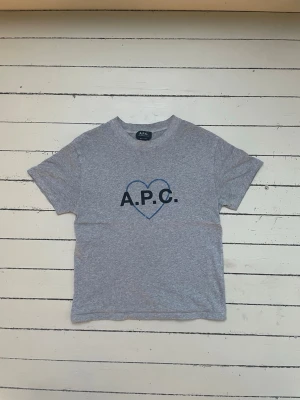 Grå A.P.C t-shirt  - Snygg grå t-shirt från A.P.C. med ett blått hjärta och svart logga på bröstet. Klassisk passform med rund halsringning och korta ärmar. Perfekt till jeans eller shorts för en avslappnad look.