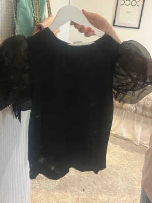 Svart topp med puffärmar - Snygg svart topp med transparenta puffärmar i mesh som ger en trendig look. Kroppsdelen är i mjukt tyg och toppen har rund halsringning. Perfekt för dig som vill sticka ut lite extra med coola detaljer.