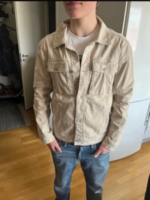 Beige jeansjacka med fickor - Snygg beige jeansjacka med klassisk krage och två bröstfickor med lock och knapp. Jackan har raka ärmar och knäppning framtill. Perfekt att slänga på sig över en t-shirt för en avslappnad look.