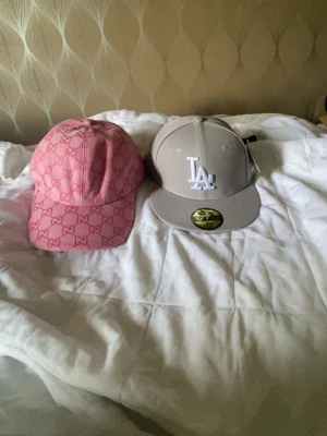 G rosa keps & LA keps - Två snygga kepsar: en rosa G-keps med GG-mönster och justerbart spänne, samt en grå New Era snapback med LA-logga i vitt framtill. Perfekta accessoarer för att lyfta din streetwear-look. De är helt nya och har aldrig använts.