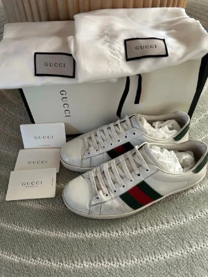Gucci Ace  - Gucci Ace sneakers i vitt läder med de ikoniska gröna och röda ränderna på sidorna. Klassisk låg modell med vita skosnören och diskret Gucci-detalj på hälen. Snygga och tidlösa sneakers som passar till allt. Levereras med originalpåse och kort.