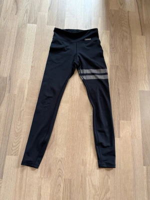 Svarta leggings från Stronger - Snygga svarta leggings från Stronger med två reflekterande ränder på ena benet. Hög midja och stretchigt material som sitter skönt. Perfekta för träning eller chill. Logga på höften och tight passform. Liten defekt på trycket