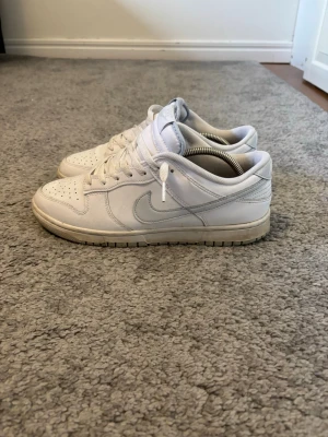 Nike Dunk Low vita sneakers - Fräscha Nike Dunk Low sneakers i helvitt läder med klassisk swoosh på sidan. Skorna har perforerad tåbox, vita skosnören och en låg siluett som ger en clean och stilren look. Perfekta för dig som gillar streetwear och vill ha en tidlös sneaker i garderoben. Allt är med, finns en liten fläck på framsidan men går att ta bort!
