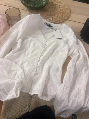 Vit blus Bik Bok - Superfin vit blus från Bik Bok med broderade blommor, v-ringning och klädda knappar framtill. Blusen har långa puffärmar med resår vid ärmslut och en lätt figursydd passform. Perfekt för en trendig och romantisk look. Storlek M. Aldrig använd lappar kvar.