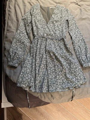 Blommig kortklänning  - Supersöt grön kortklänning från Glamorous med vitt blommönster, v-ringning och långa ärmar. Klänningen har knytband i midjan och dragkedja bak. Perfekt för vår och sommar!