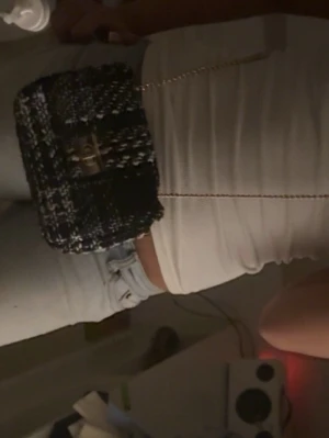 Svartvit axelväska med kedja - Trendig axelväska i svartvitt tweedtyg med guldfärgad kedjerem och snyggt vridlås framtill. Väskan har en kompakt, rektangulär form och detaljer i metall. Perfekt för dig som gillar en stilren och modern look.