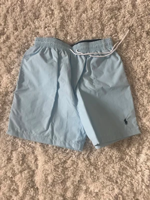Ljusblå badbyxor från Polo Ralph Lauren - Snygga ljusblå badbyxor från Polo Ralph Lauren med klassisk logga broderad i mörkblått på benet. Modellen har elastisk midja med vit dragsko för skön passform. Tillverkade i snabbtorkande material, perfekta för stranden eller poolen. Skriv om du har frågor!
