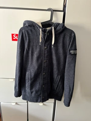 Mörkgrå hoodie från Abercrombie - Mörkgrå hoodie från Abercrombie & Fitch med dragkedja och knappar framtill, vit dragsko i huvan och två fickor. Storlek XS, jag är 168 och den sitter lite för stort. Pris kan självklart diskuteras 