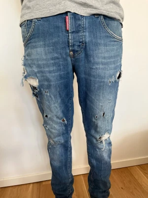 Blå slitna jeans från Dsquared2 - Säljer ett par blå jeans från Dsquared2 med tydliga slitningar och hål för en trendig, avslappnad look. Jeansen har klassisk femficksmodell och röd logotagg vid fickan. Perfekta för dig som gillar streetstyle och vill sticka ut.