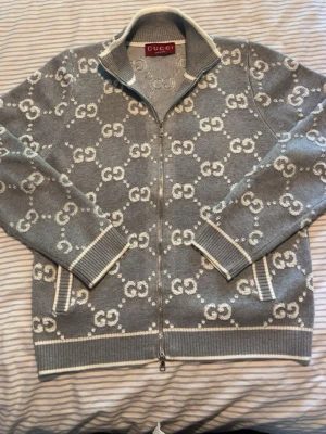 Grå ziptröja med GG-mönster från Gucci - Snygg grå ziptröja från Gucci med hel dragkedja och ikoniskt GG-mönster i vitt över hela tröjan. Tröjan har ribbade muddar, ståkrage och vita detaljer vid fickor och kanter. Perfekt för dig som gillar exklusiv streetstyle. Den är väldigt snygg och perfekt inför sommaren. Den är i storlek M. Vid några funderingar är det bara att höra av sig☺️