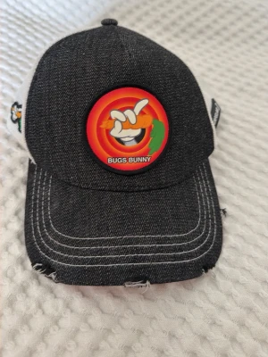 Svart Bugs Bunny keps med brodyr - Säljer en svart keps med vit mesh baktill och broderad Bugs Bunny-detalj framtill. Kepsen har en cool patch med Bugs Bunny-logga och morot, samt broderad morot på sidan. Justerbar baktill och med slitna detaljer på skärmen för en trendig look.